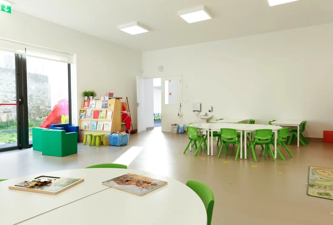 Sala do jardim de infância, luminosa, com mesas brancas e cadeiras verdes abertas para o recreio